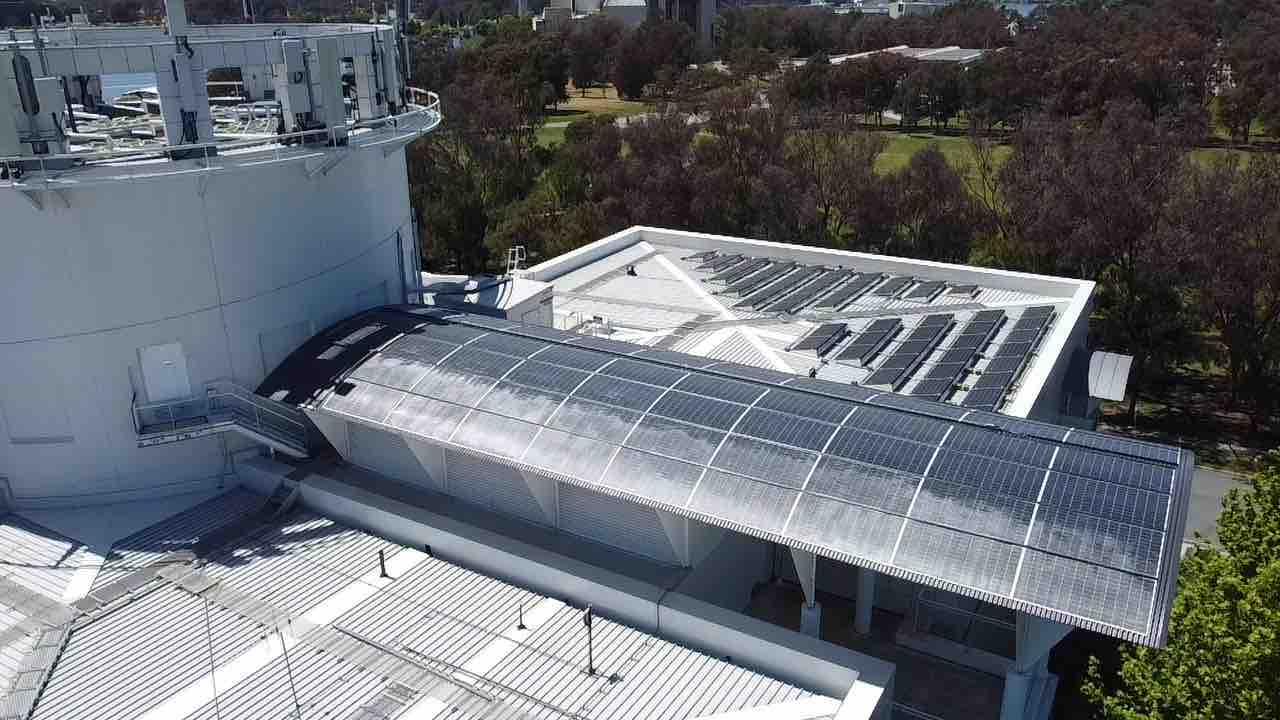 Aerial view of the white Questacon building with a curved solar canopy and a tilted array on the flat roof beyond