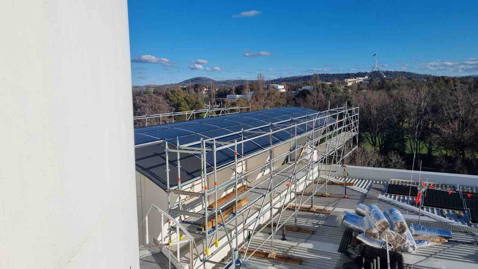 Curved rooftop solar array under scaffold, with the Parliament House flagpole on the Canberra horizon