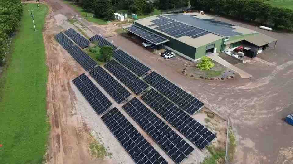 Aerial of a Northern Territory agricultural site: rows of ground mount solar on red earth beside a green shed carrying a full rooftop array and a carport